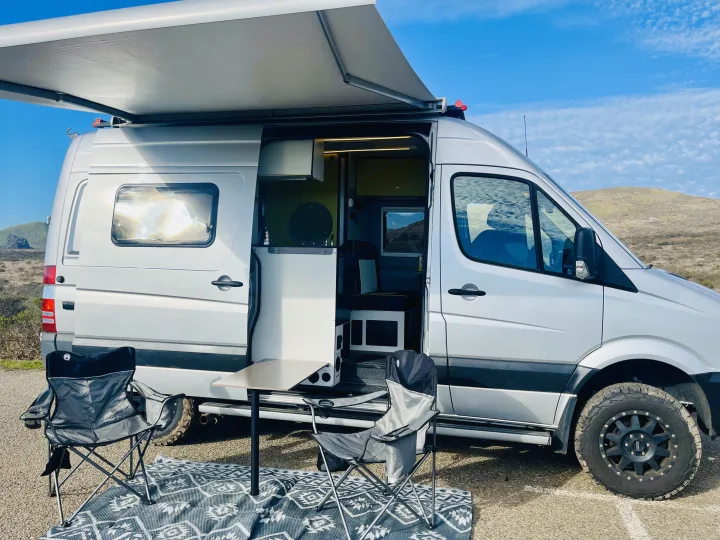 This rv rental features a silver camper van with an extended awning, open side door, outdoor chairs, and table setup for camping.. Winnebago Revel 2019