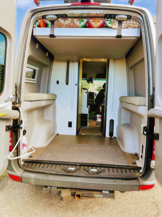 View into an RV rental's rear, revealing an upper bed, side benches, wood floor, and front cab area.. Winnebago Revel 2019