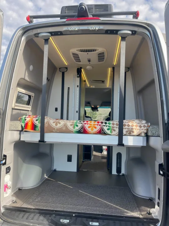 Inside a modern rv rental camper van, revealing a raised bed, ceiling AC, and warm strip lighting.. Winnebago Revel 2019