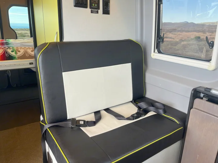 Inside an RV rental: a black and white bench seat with seatbelts. Windows reveal ocean views on one side and hills on the other.. Winnebago Revel 2019