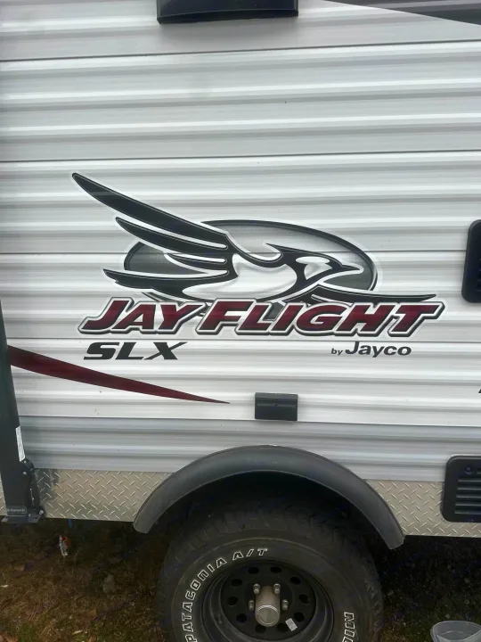 Side view of a white Jayco Jay Flight SLX travel trailer, ready for your RV rental. Features ribbed siding, logo, and a tire.. Jayco Jay Flight SLX 2015