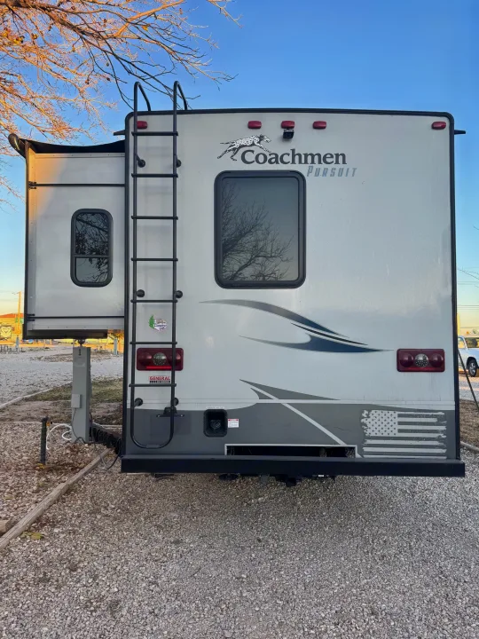 Rear view of a white Coachmen Pursuit RV rental parked on gravel, showing a slide-out, ladder, and American flag decal.. Forest River Coachmen Pursuit 2020