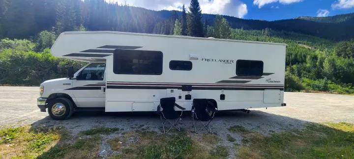 White Coachmen Freelander RV rental parked amid green mountains and forest. Two chairs await relaxation by this motorhome.. Freelander Coachman 2023