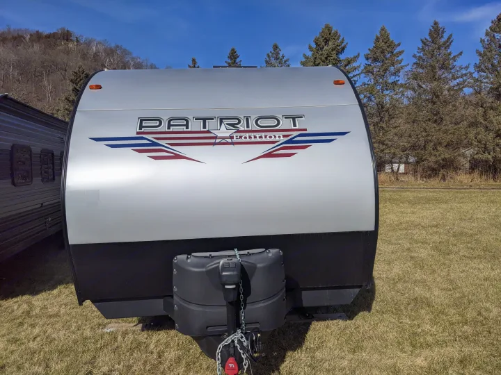 A silver Patriot Edition travel trailer, perfect for your next rv rental. Shows front with hitch & bright sky.. Forest River Patriot 26DBH 2022