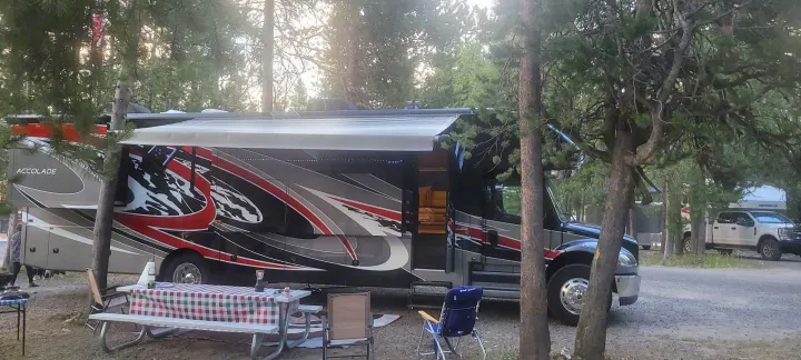 An Accolade RV rental is set up for camping, with an awning, picnic table, and chairs in a wooded campsite.. Entegra Coach Accolade XL 2021