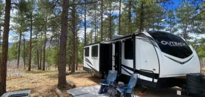 Spacious RV rental parked in a peaceful pine forest campsite. Awning extended, chairs out for relaxation.. Keystone RV Outback Ultra-Lite 2020