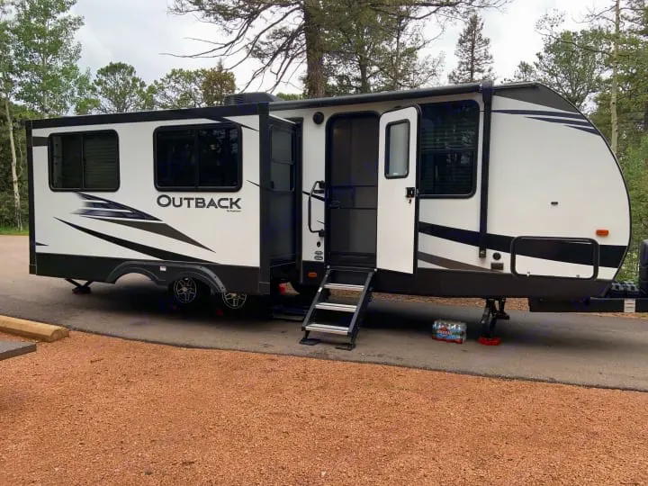 This white Outback travel trailer, an RV rental, is set up at a wooded campsite with steps extended and awning out.. Keystone RV Outback Ultra-Lite 2020