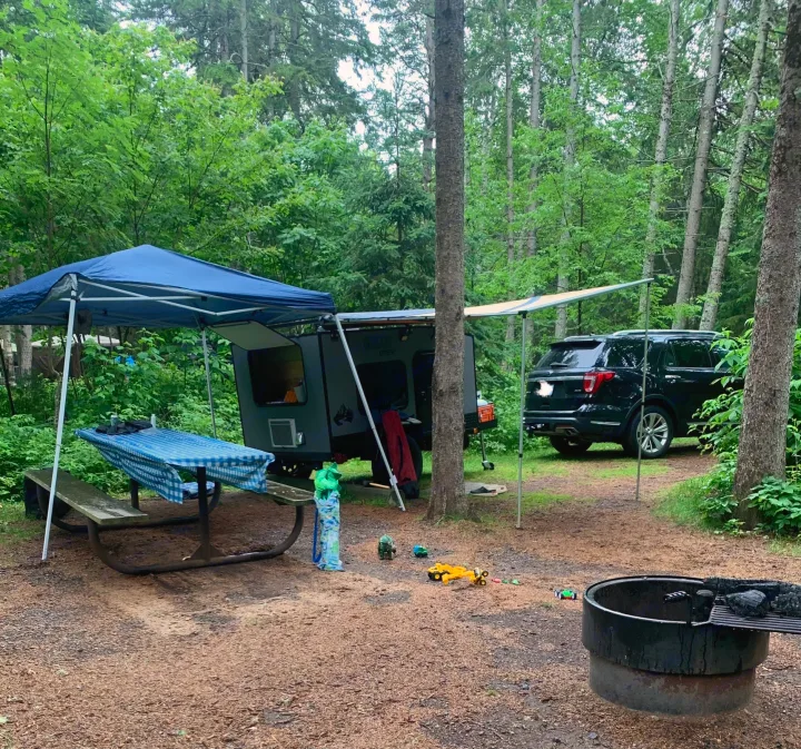 Family camping at a wooded campsite featuring an RV rental (teardrop camper), SUV, covered picnic table, and a fire pit.. Outbound Extreme 2021