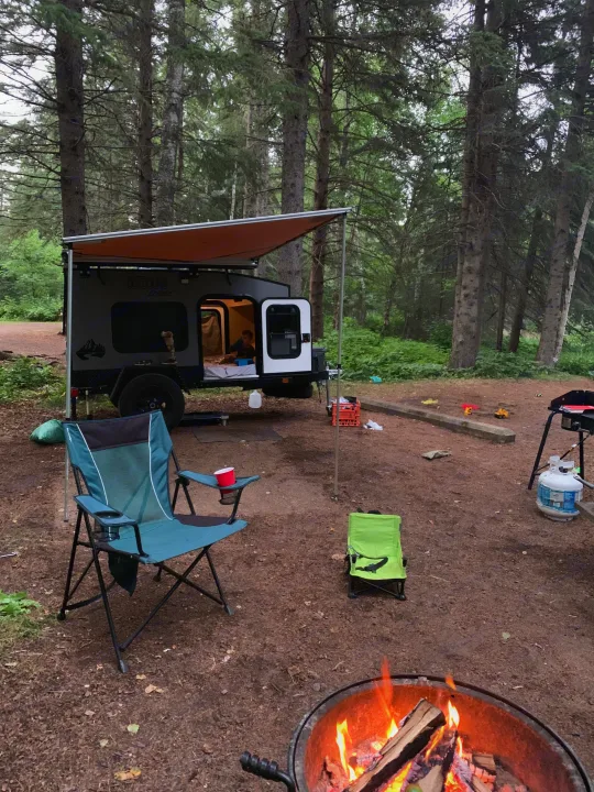 Forest campsite scene with an RV rental, featuring a child inside the camper, a glowing campfire, and camping chairs.. Outbound Extreme 2021