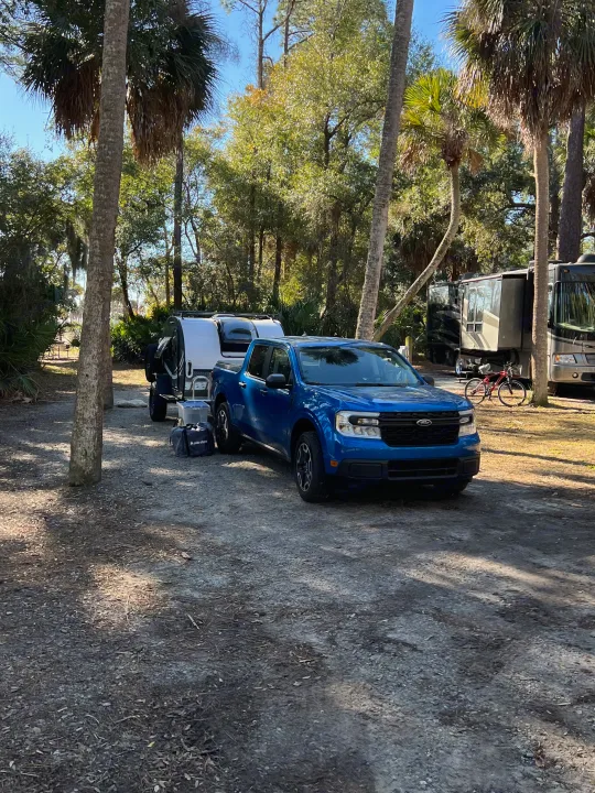 Blue truck with rv rental camper at a sunny campsite. Larger motorhomes and palm trees in this serene camping setting.. nuCamp T@G 2023