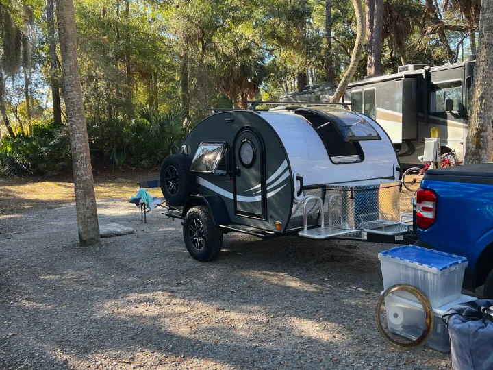 Compact teardrop travel trailer and blue pickup truck at a shaded campground, ideal for an rv rental getaway.. nuCamp T@G 2023