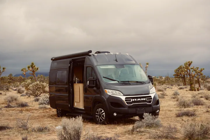 Dark grey Ram Promaster RV rental camper van parked in a desert with Joshua trees under an overcast sky, side door open.. Noovo Lite 2023