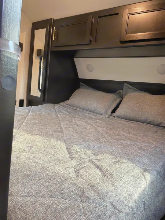 Cozy RV rental bedroom featuring gray bedding, dark overhead cabinets, and a built-in mirror for a comfortable sleeping area.. Forest River Patriot 26DBH 2022