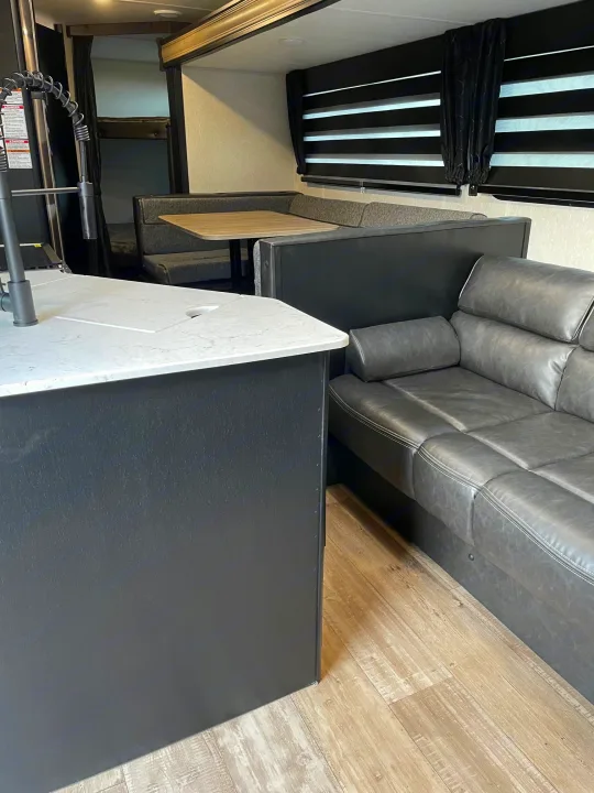 Modern rv rental interior with a kitchen island, grey sofa, dinette, and visible bunk beds. Spacious and functional design.. Forest River Patriot 26DBH 2022