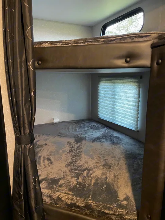 An RV rental's bunkhouse with dark curtains, a top bunk window, and a lower bunk with window blinds.. Forest River Patriot 26DBH 2022