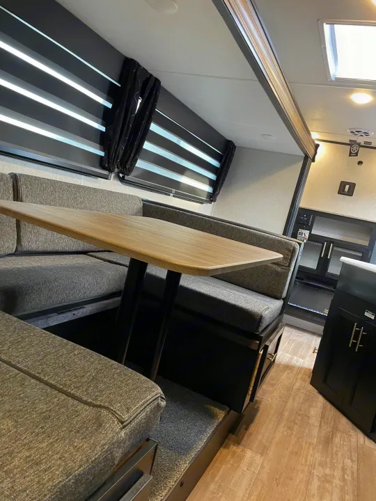 Modern RV rental interior: a cozy dinette with gray fabric seating, light wood table, and sleek window blinds.. Forest River Patriot 26DBH 2022