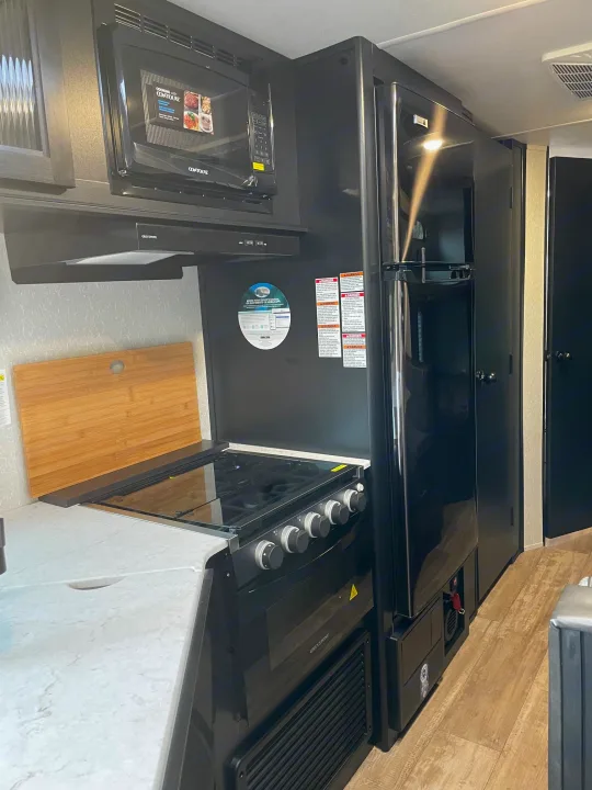 Inside an RV rental, a sleek black kitchen features a microwave, refrigerator, stove, and light countertop.. Forest River Patriot 26DBH 2022