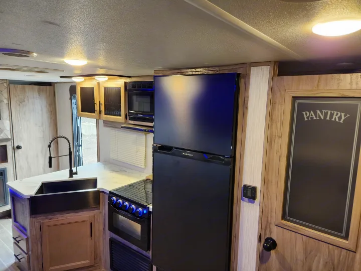 Spacious modern RV rental kitchen featuring a black Furrion fridge, stove, microwave, sink, and "Pantry" door.. Forest River Cherokee Alpha Wolf 2020