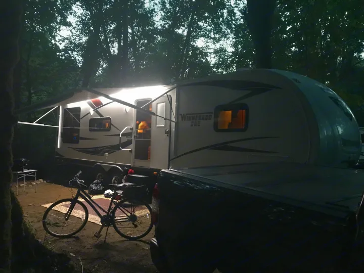 An RV rental illuminated at a dark campsite, featuring an extended awning, a parked truck, and a bicycle.. Keystone RV Passport Grand Touring 2014