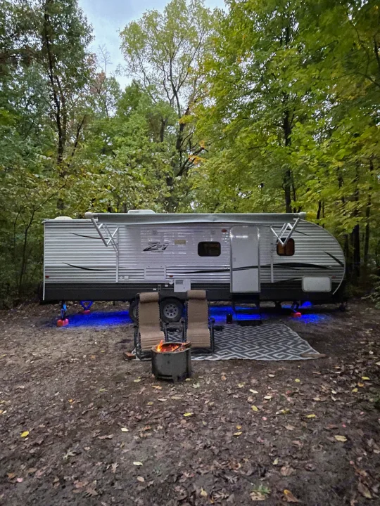 Cozy RV rental campsite at dusk. Blue lights glow under the camper, with chairs & a fire pit ready for relaxation.. Crossroads RV Z-1 2017