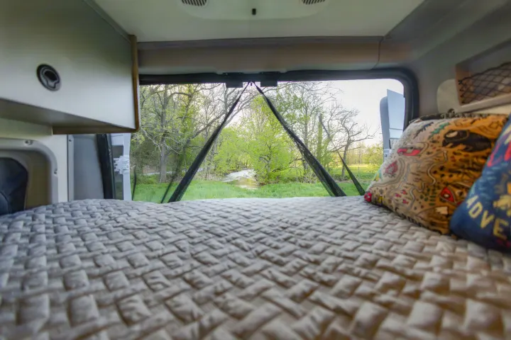 RV rental: Comfy bed, pillows, and open back doors reveal a serene view of a stream flowing through lush green trees.. Winnebago Solis Pocket 2022