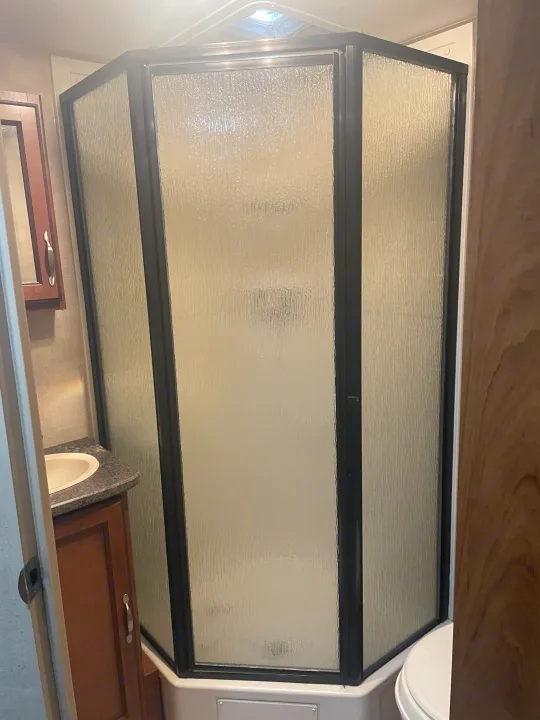 RV rental bathroom features a compact shower with frosted glass and dark frames, a sink with a wood vanity.. Keystone RV Passport Grand Touring 2014