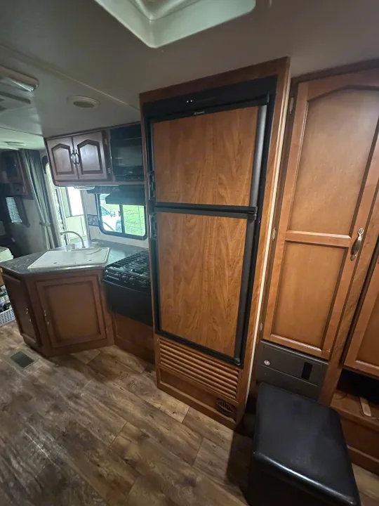 Inside an RV rental, a compact kitchen features a wood-paneled fridge, sink, and gas stove on laminate floors.. Keystone RV Passport Grand Touring 2014
