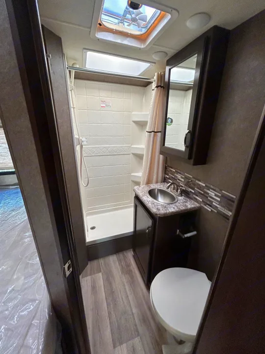 Compact RV rental bathroom with shower, toilet, and sink vanity. Features wood-look floor, overhead skylight, and mirror.. Thor Freedom Elite 22HE 2020