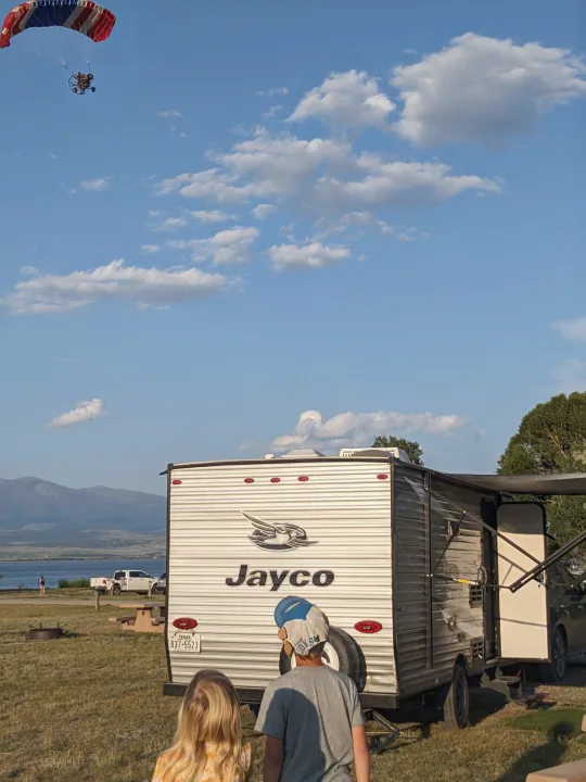 Kids watch a paramotor over an RV rental by a lake & mountains. Ideal for outdoor family adventures!. Jayco Jay Flight SLX 2020