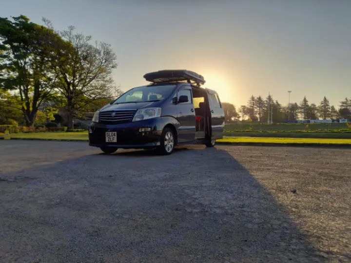 Explore with this dark blue campervan RV rental, featuring a roof box and open side door, parked at sunset on gravel.. Toyota Alphard 2024