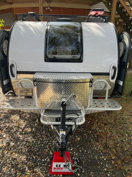 Front view of a white teardrop travel trailer on gravel, showing its storage box, hitch, and front window. Perfect for RV rental.. nuCamp T@G 2023