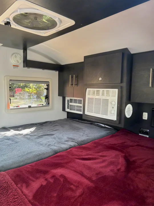 RV rental interior featuring two comfortable beds, a window with outdoor views, overhead cabinets, AC, and a ceiling fan.. nuCamp T@G 2023