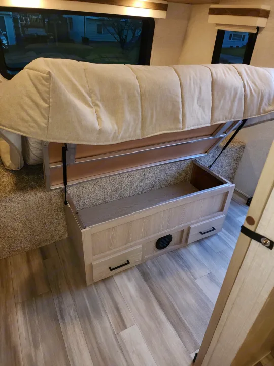 This RV rental interior features a lifted bed with gas struts for under-bed storage, a wood-toned storage bench with drawers, and a light wood-look floor.. Forest River Rockwood Ultra Lite 2022