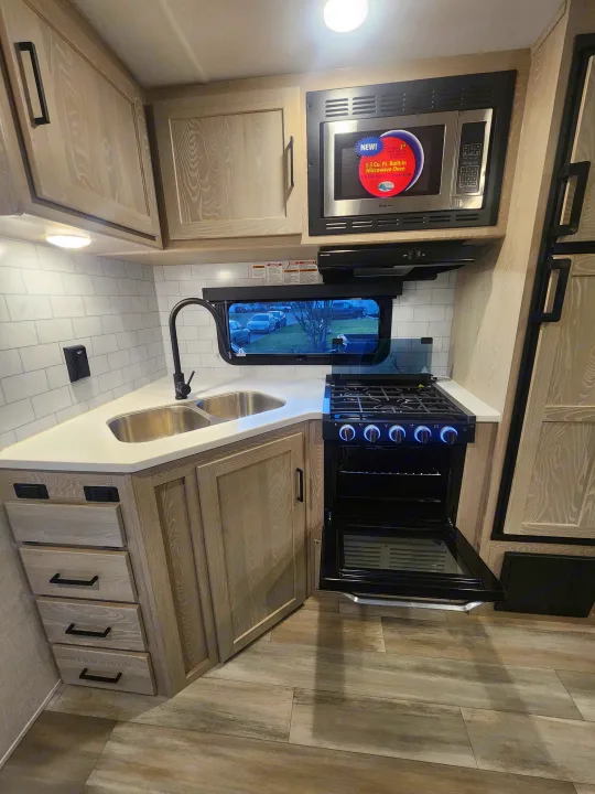 This compact RV rental kitchen features light wood cabinets, white countertops, a sink, microwave, and an open oven.. Forest River Rockwood Ultra Lite 2022