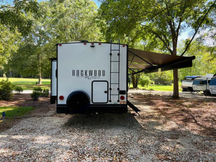 White Rockwood RV rental, with awning extended, parked on gravel in a shaded campsite. Perfect for outdoor adventures.. Forest River Rockwood Ultra Lite 2022