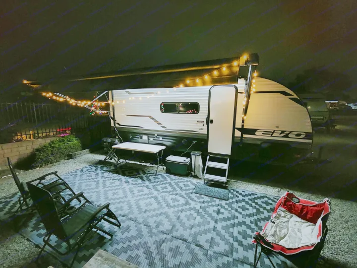 Cozy RV rental campsite at night, featuring string lights under the awning, an outdoor rug, chairs, and a table setup.. Other Other 2024