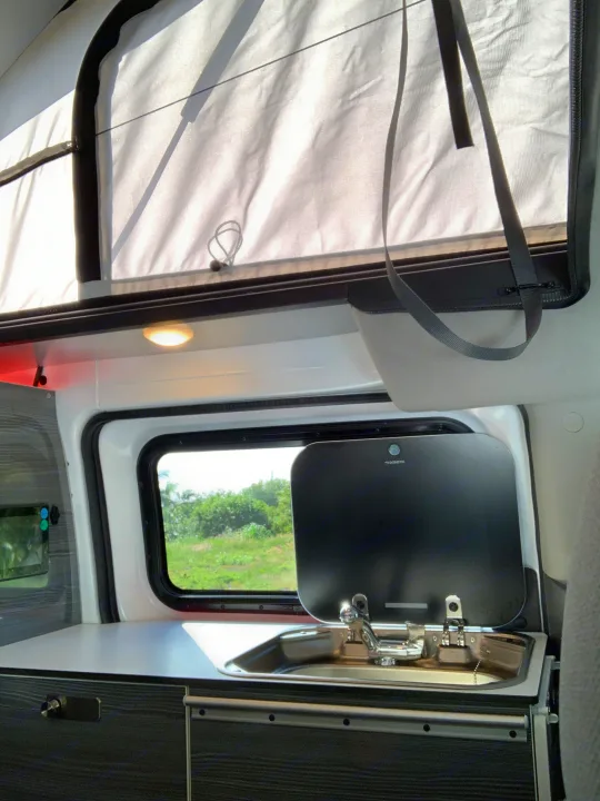 Compact **RV rental** interior: a sleek kitchen features a stainless sink with Dometic cover, under a bright pop-top roof. Enjoy the view from your **campervan**.. NIssan Envy 2021