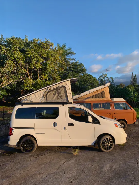 Two pop-top camper vans, a white and an orange rv rental, parked among lush trees. Ideal for outdoor travel and adventure.. NIssan Envy 2021