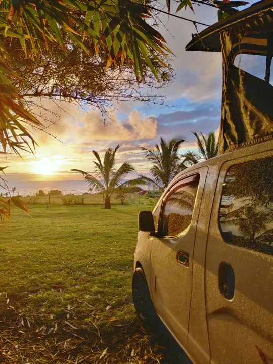 White RV rental, covered in dew, parked on a grassy field at sunrise, overlooking the ocean and palm trees.. NIssan Envy 2021