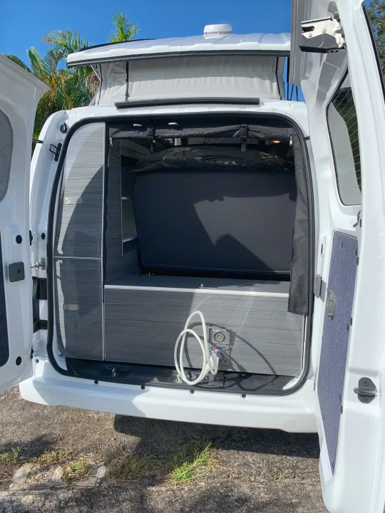 RV rental: Rear interior of a white pop-top camper van with doors open, showing a bed, grey storage, and outdoor shower hookup.. NIssan Envy 2021