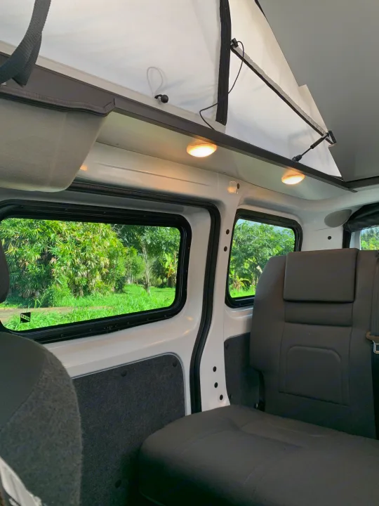 Cozy RV rental interior: pop-top roof, comfortable seating, and large windows revealing lush green nature outside. Ideal for travel.. NIssan Envy 2021
