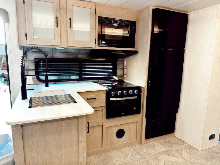 Compact RV rental kitchen features light wood cabinets, black appliances (fridge, microwave, stove), and a modern sink.. Forest River Wildwood X-Lite Platinum 2024