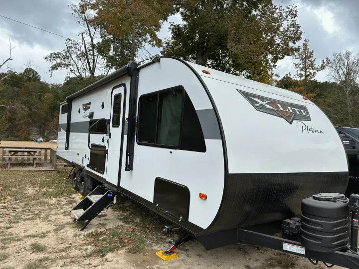 White & black rv rental travel trailer set up at a scenic campsite, with steps down & picnic area nearby.. Forest River Wildwood X-Lite Platinum 2024