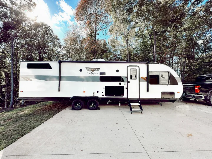 White X-Lite Platinum RV rental travel trailer parked on a concrete driveway, hooked to a black pickup truck. Trees in background.. Forest River Wildwood X-Lite Platinum 2024