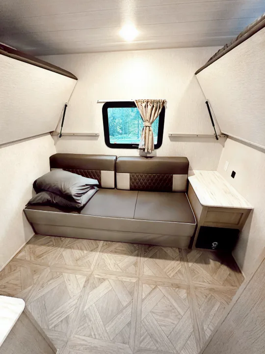 Cozy A-frame RV rental interior with a convertible sofa, small side table, and window view of trees. Ideal for compact travel.. Forest River Wildwood X-Lite Platinum 2024