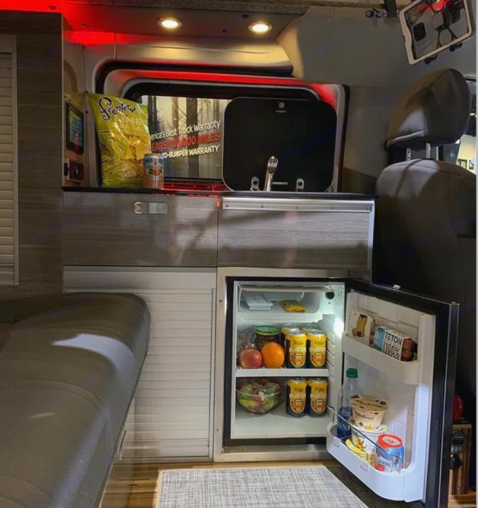 Explore an RV rental's interior: a compact camper kitchen with a stocked fridge, sink, and seating area. Perfect for mobile living.. NIssan Envy 2021