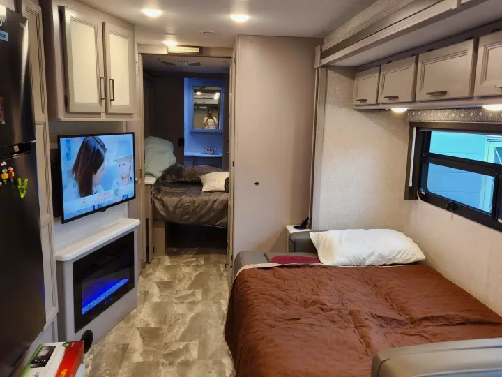 Modern RV rental interior featuring a main bed, TV with fireplace, refrigerator, and a glimpse of the rear bedroom/bathroom.. Thor Windsport 2023