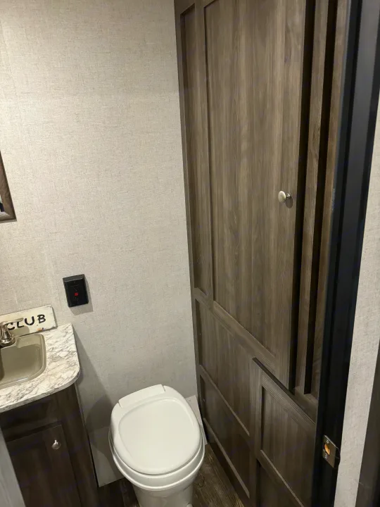 RV rental bathroom with white toilet, sink, dark wood cabinetry, and a GFCI outlet. Walls are light textured grey.. Starcraft Autumn Ridge Outfitter 2020