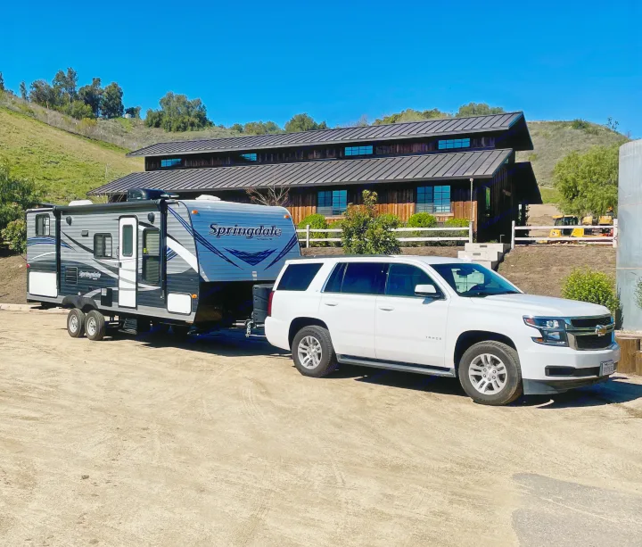 White SUV towing a large Springdale travel trailer, perfect for an RV rental getaway. Parked on a dirt lot, with a building and hills.. Keystone RV Springdale Eco-Lite 2018