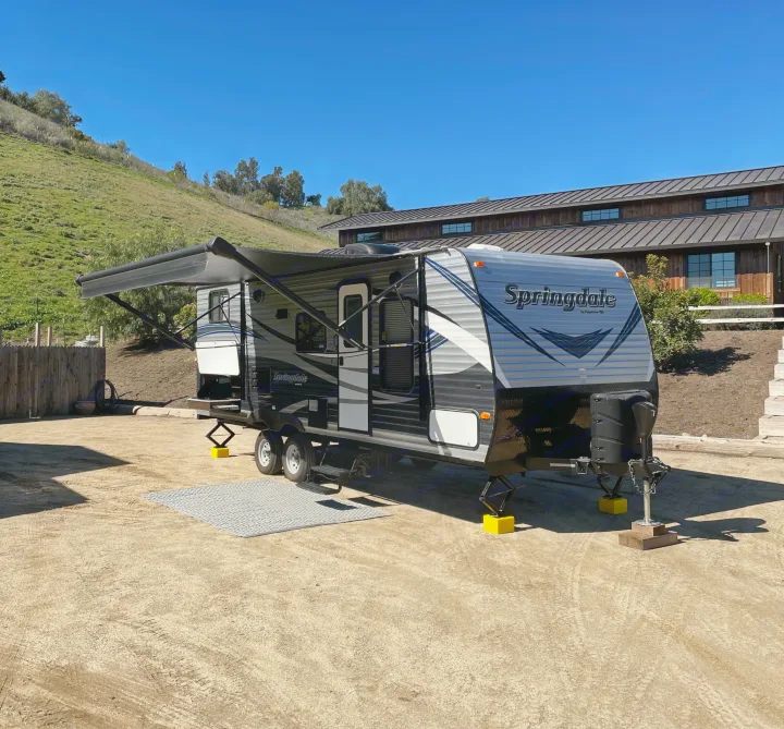 A grey 'Springdale' travel trailer RV rental, with awning out, parked on a dirt lot. Yellow leveling blocks are visible.. Keystone RV Springdale Eco-Lite 2018